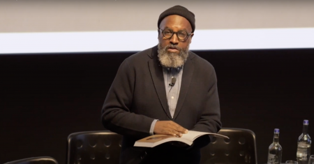 Man standing on stage reading poetry from a book in his hand